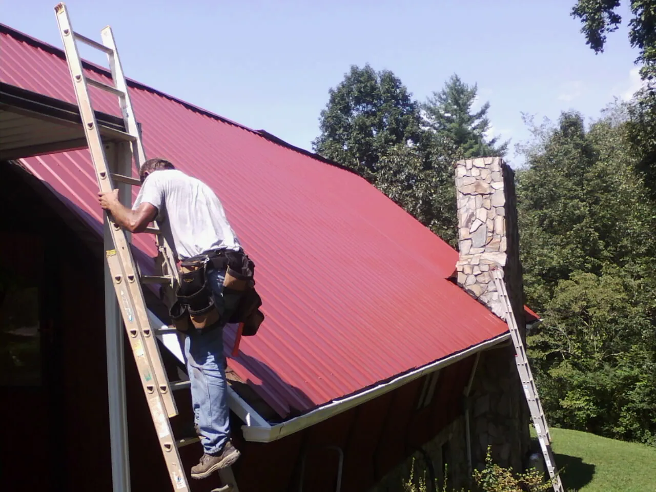 Roofer on a ladder beside a red metal roof and stone chimney — Soffit & Fascia Repair by Roofing Brothers in Victory Mills